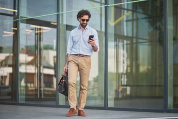 A well-dressed individual in a light blue shirt and tan pants is walking outside a contemporary...