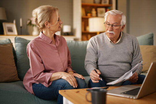 An older couple sits on a couch in their living room during the day, reviewing documents and using a laptop. They appear to be planning their finances, possibly discussing life insurance.