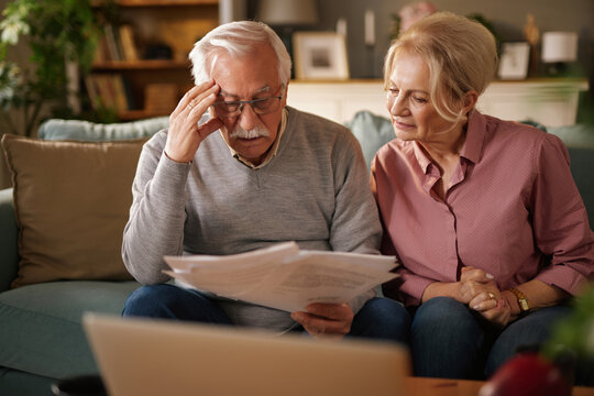 An older Caucasian couple sits on a couch in their living room during the daytime. The man holds documents, looking stressed. The woman watches him with a concerned expression. - Powered by Adobe