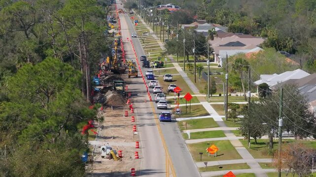 Street construction in Florida suburb with cars navigating single-lane traffic past construction vehicles, trenching machines and utility signs.