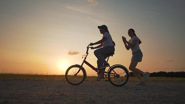 Girl learns to ride bike outdoors. Child pedaling at sunset while mother runs behind offering help. Outdoor support and encouragement during training moment with bike and happy child presence