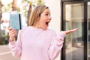 Young blonde woman holding a passport at outdoors with surprise facial expression