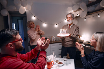 Elderly man holding birthday cake with lit candles surrounded by family members clapping in warmly...