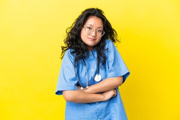 Young surgeon doctor asian woman isolated on yellow background making doubts gesture while lifting...