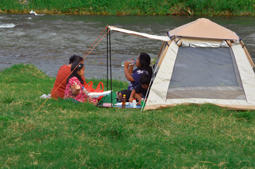 A mother refreshes herself with bottled water while children and another woman sit nearby enjoying...