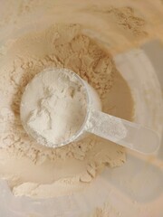 A transparent scoop filled with fine, beige protein powder resting on more powder inside a container