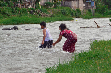 A young boy confidently crosses the flowing river stream, soon followed by a young girl who carefully makes her way through the water