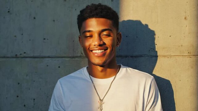 A young African man with short curly hair smiles, showcasing his dimples. He wears a white t-shirt and a necklace, standing against a concrete wall..