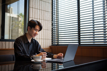 Fototapeta premium Smiling Asian entrepreneur using computer and taking notes in a modern office.