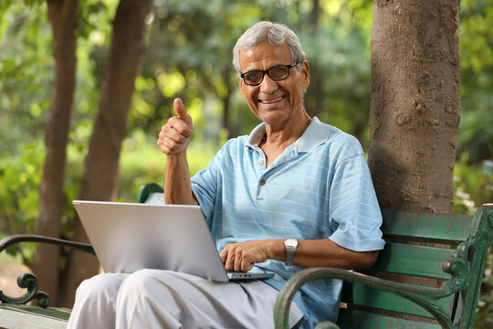 Indian Happy senior man working on laptop and showing thumbs up while sitting on the bench at the park.