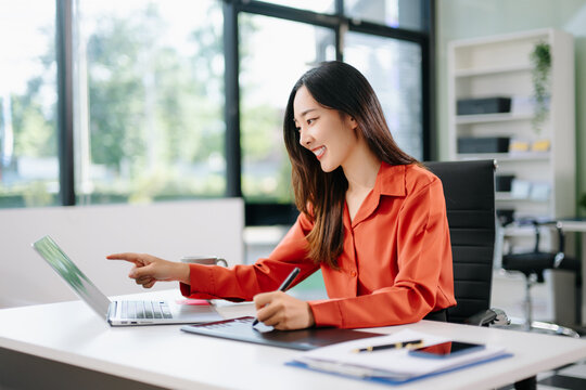 Happy Asian woman creating digital content on laptop and drawing tablet in modern office.