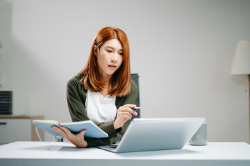 Young Asian woman in office focusing on laptop and writing in notebook. Concept of productivity,...