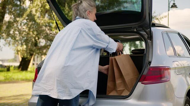 Older Caucasian mature business woman shopper customer senior buyer client female elderly lady car auto driver opening automobile trunk put loading shopping bags in vehicle delivery order in park city