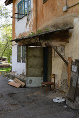Old entrance with broken door, weathered chair, peeling wall, and overgrown roof in urban area