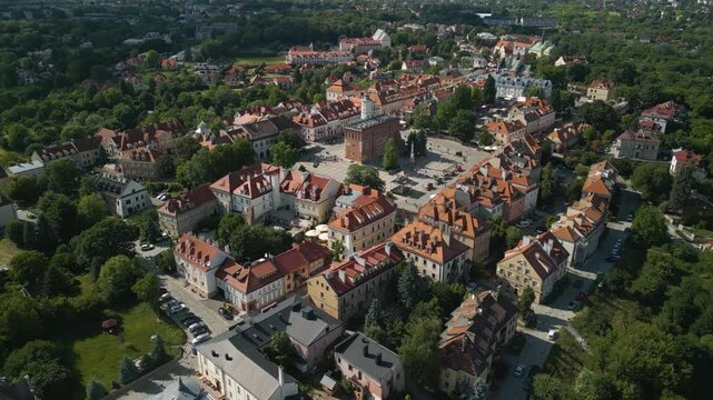 Aerial panoramic view of Sandomierz Old Town in Poland with colorful historic buildings, a central market square, and red rooftops surrounded by greenery on a sunny summer day.