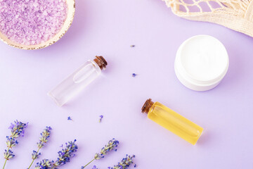 Lavender bath salt in bowl, cream jar, lavender flowers, essential oils on purple background. Natural herbal medicine, aromatherapy concept. Top view, flat lay