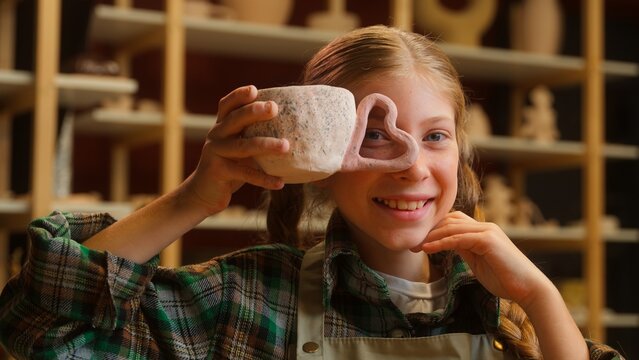 Creative talented Caucasian potter little girl schoolgirl student child kid artist making clay craft cup mug heart shape pottery artwork having fun creation looking at camera indoors workshop studio