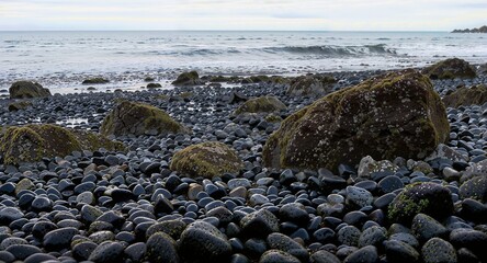 Rocky beach with smooth pebbles and large stones. The ocean waves gently lap at the shore under a cloudy sky. A serene coastal landscape.