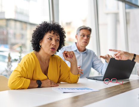 A group of businesspeople are gathered around a table, discussing something. Scene is serious and focused, as the group appears to be engaged in a discussion or decision-making process. There is a lot - Powered by Adobe