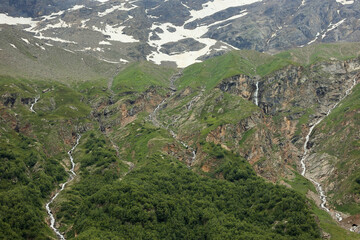 View Of Steep Mountain Slopes With Patches Of Green Vegetation, Rocky Outcrops, Snow Remnants, And...