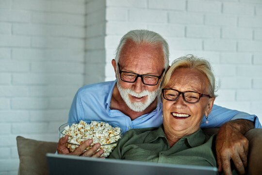 Portrait of an elderly senior happy couple using laptop at night, using modern technology having fun with glowing screen in dark office or at home, watching tv together eating popcorn at home. Shot of - Powered by Adobe
