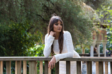 Naklejka premium Young and beautiful brunette woman in white knitted dress, makes different expressions leaning on the wooden railing of a bridge in the park. In the background trees and vegetation.