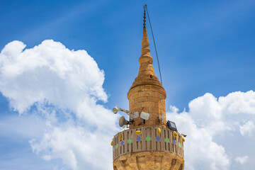 View of the top of a minaret with loudspeakers and CCTV cameras against a blue sky with white fluffy cumulus clouds