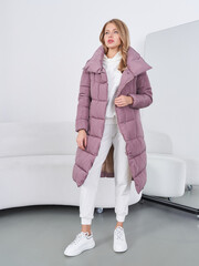 Woman wearing a long, mauve quilted coat over a white outfit, posing in a modern room with white...