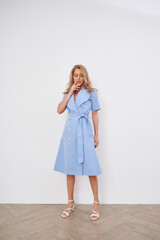 Elegant woman in light blue belted dress and white heels stands on wooden floor, hand gently touching lips, exuding sophistication and style in fashion-forward attire