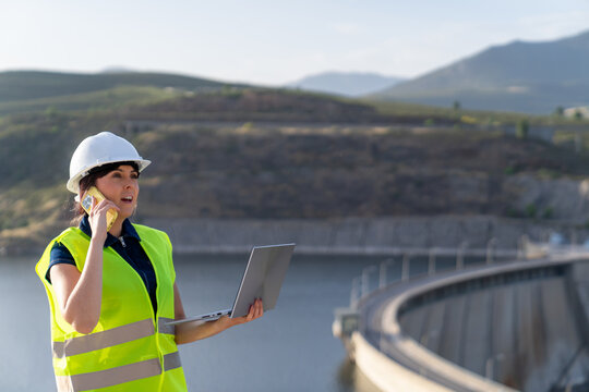 Female engineer using laptop and talking on phone at dam