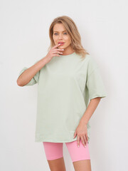 Young woman with blonde hair wearing a light green oversized T-shirt and pink shorts, posed against a neutral background. She touches her face thoughtfully, with a relaxed expression
