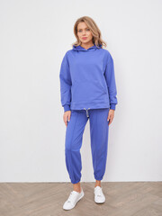 Woman in a blue hoodie and jogger pants stands confidently, wearing white sneakers. Her hands are...