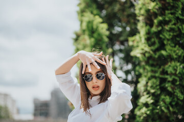 A fashionable young woman wearing sunglasses and posing confidently outdoors near greenery. The...