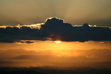 Scenic sunset with the sun peeking through a dark cloud. The sky is bathed in warm golden hues, while the illuminated cloud edges add depth and contrast to the image.