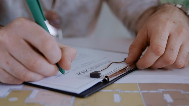 Handheld close-up shot showing an anonymous man signing a mortgage contract while detailed blueprints of a flat lay beside him on the table.