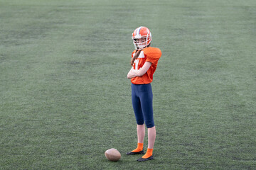 A woman dressed in orange football gear poses confidently on a green field. It is during practice,...
