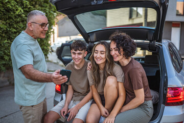 father show mobile phone screen to his family who sit in car trunk