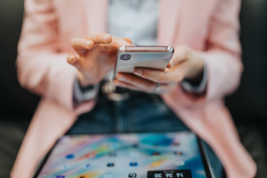 Person wearing pink jacket interacts with a mobile phone while multitasking. Tablet with colorful screen visible in foreground, emphasizing connectivity and contemporary digital technology.