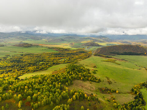 A sweeping aerial view of an expansive autumn landscape featuring vibrant yellow and green forests, rolling hills, and distant mountains