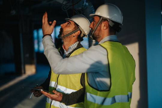 Two construction supervisors wearing safety vests and helmets inspecting a building site with intent. The scene is lit with natural and indoor lighting, highlighting their professional activity.