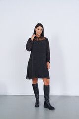 A woman stands confidently, wearing a black long-sleeve dress with small polka dots and knee-high leather boots. Her dark hair is straight, accentuating the chic modern outfit