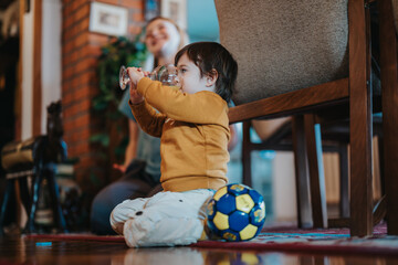 A toddler drinks from a glass while sitting on a rug with a toy soccer ball nearby, depicting a...