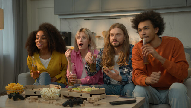 Four diversity multiethnic friends Caucasian female women girls African American male guys men enjoying watching TV television sport game program at home couch eating pizza amazed weekend friendship