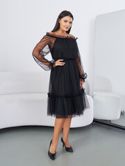Woman in elegant black dress with sheer sleeves stands smiling in room beside white sofa and large...
