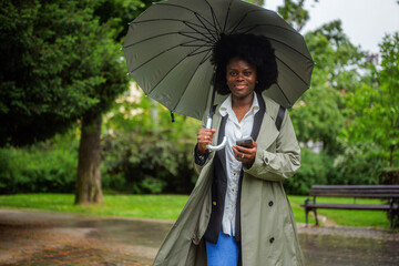 Businesswoman walking through a city park, holding an umbrella and checking her smartphone during a...