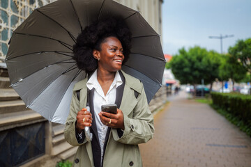 Cheerful manager walking confidently along a city street, holding an umbrella in one hand and a smartphone in the other, smiling brightly while navigating the rainy commute to work