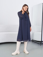 Woman in navy polka dot dress touches her hair, standing in white knee-high boots. She is near a large mirror and a curved white couch. Her gaze is gentle and thoughtful