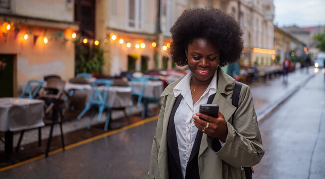 Happy businesswoman wearing a trench coat and carrying a backpack is using her smartphone while walking near an outdoors restaurant in the city during a cloudy day - Powered by Adobe