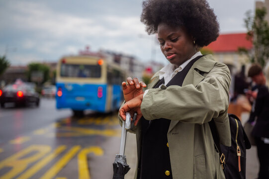 Businesswoman is checking the time on her smartwatch while waiting for the bus at a bus stop on a rainy day, holding an umbrella and wearing a trench coat