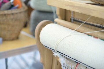 Handwoven textile rolled onto the beam of a wooden loom, with yarn threads passing through the...